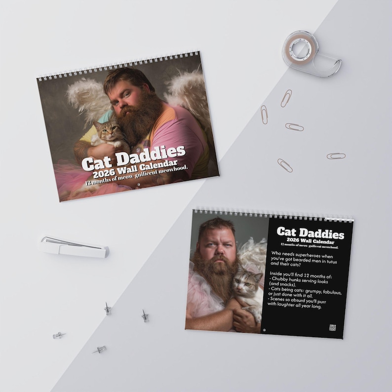 May include: A spiral-bound wall calendar titled "Cat Daddies 2026." The calendar features a man with a beard and a cat. The text on the calendar reads "12 months of meow-nificent meowhood." A second calendar is also visible.