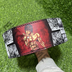 May include: A red, black and white weightlifting belt featuring a graphic of a muscular anime character. The character is shown in a fighting pose against a red background.
