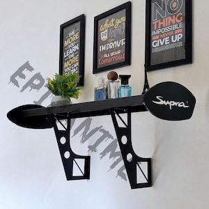 May include: A black wall-mounted shelf designed to resemble a car spoiler, featuring two side end plates with the word Supra printed in white. The shelf is supported by two angular metal brackets with circular cutouts and holds a small potted plant.