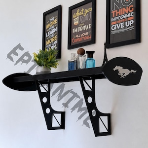 May include: A black wall-mounted shelf designed to resemble a car spoiler, featuring two vertical support brackets with circular cutouts and a silver-toned horse emblem on the right side. The shelf holds a small potted plant and glass bottles.