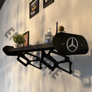 May include: Black wall-mounted shelf with a car spoiler design. The shelf features a black finish with a Mercedes-Benz logo on the side. Decorative items, including a small plant and perfume bottles, are displayed on the shelf.
