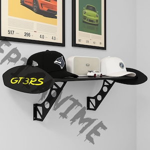 May include: A black shelf with a car spoiler design, holding a black hat with a wolf logo, a white hat with a blue emblem, a white speaker, and a small toy car. Two framed car posters are above the shelf.