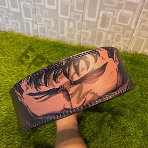 K&ouml;nnte beinhalten: Schwarzer Gewichtheberg&uuml;rtel mit einer detaillierten Illustration eines Gesichts mit rotem Auge. Der G&uuml;rtel ist aus schwarzem Leder gefertigt und hat gen&auml;hte Kanten. Das Design ist auf der Vorderseite aufgedruckt.