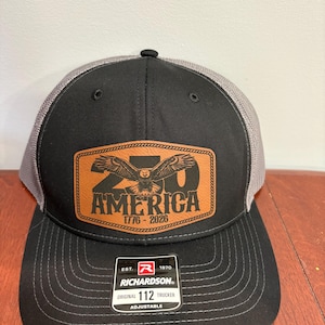 May include: Black and gray trucker hat with a leather patch featuring an eagle and the text "20 AMERICA 1776-2026". The hat has a curved brim and an adjustable strap. The brand "Richardson" is visible.