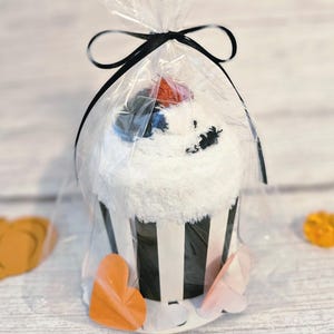May include: A cupcake-shaped item made of white fuzzy fabric inside a clear plastic bag tied with a black ribbon. The base is a black and white striped paper wrapper. A small orange accent sits on top, and orange heart-shaped cutouts are at the base.