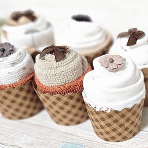 May include: A collection of six cupcake-shaped baby washcloths. Each cupcake has a white, cream, or tan 'frosting' and sits in a brown checkered paper cup. Some are topped with decorative flowers or bows.