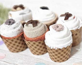 May include: A collection of six cupcake-shaped baby washcloths. Each cupcake has a white, cream, or tan 'frosting' and sits in a brown checkered paper cup. Some are topped with decorative flowers or bows.