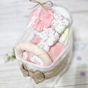 May include: A pink and white baby gift basket containing a pink and white blanket, a pink and white towel, and a pink and white hat. The basket is wrapped in clear plastic and tied with a brown ribbon. The basket has a white polka dot pattern and the words "little love" embroidered on it.