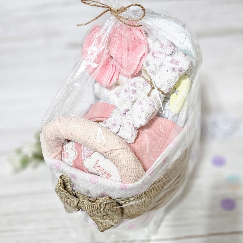 May include: A pink and white baby gift basket containing a pink and white blanket, a pink and white towel, and a pink and white hat. The basket is wrapped in clear plastic and tied with a brown ribbon. The basket has a white polka dot pattern and the words "little love" embroidered on it.