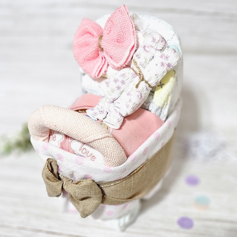 May include: A pink and white nappy cake with a pink bow on top. The cake is decorated with a burlap ribbon and a pink and white polka dot fabric. The cake is sitting on a white surface.