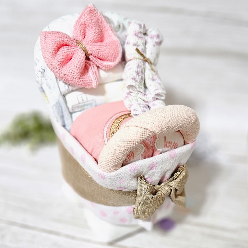 May include: A pink and white baby shower gift basket with a pink bow, a pink and white blanket, and a pink and white stuffed toy. The basket is decorated with pink polka dots and a gold bow.
