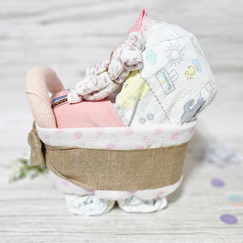 May include: A pink and white nappy cake with a brown hessian ribbon. The cake is decorated with a pink and white polka dot pattern and a white bow. The cake is shaped like a pram and has a pink and white blanket on top.
