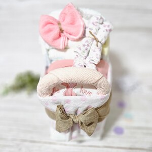 May include: A pink and white baby stroller diaper cake with a pink bow, a white and pink towel, and a white and purple patterned fabric. The stroller is decorated with a brown bow and the words "Little Love".