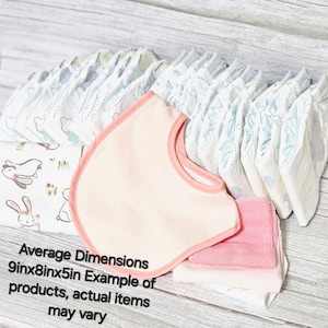 May include: A set of baby essentials including a pink bib, a stack of white and blue patterned baby nappies, and a stack of pink and white face cloths. Average Dimensions 9in x 8in x 5in. Example of products, actual items may vary.