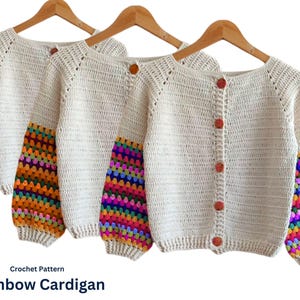 Peut inclure: Cardigan en crochet crème avec des manches arc-en-ciel, présenté sur des cintres en bois. Le cardigan a une encolure ronde et est boutonné sur le devant avec des boutons orange. Les manches présentent un motif géométrique coloré. Le texte "Crochet Pattern Rainbow Cardigan" est visible.