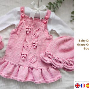 May include: A pink knitted baby dress set, featuring a dress with grape and leaf motifs, a matching hat, and booties. The dress has a frilled hem and buttoned straps. The set is presented on a white backdrop.
