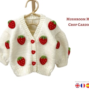 Op de afbeelding: Crèmekleurig, gebreid crop-cardigan versierd met rode aardbeienapplicaties en groene bladeren. Het vest heeft een V-hals, hartvormige knopen en de tekst "MUSHROOM MOTIF CROP CARDIGAN". Het kledingstuk wordt op een houten hanger getoond.