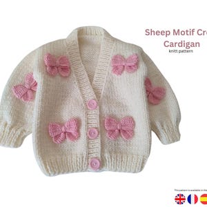 Könnte beinhalten: Cremefarbene Strickjacke mit rosa Schleifenakzenten. Der Pullover hat einen V-Ausschnitt, einen Knopfverschluss und ist mit mehreren rosa Schleifen verziert. Der Text auf dem Bild lautet "Sheep Motif Crop Cardigan knitt pattern".