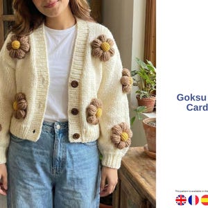 Puede incluir: Cárdigan Goksu Daisy de color crema con aplicaciones de flores marrones y amarillas. El cárdigan tiene cuello en V, mangas largas y botones marrones. El texto "Goksu Daisy Cardigan" es visible.