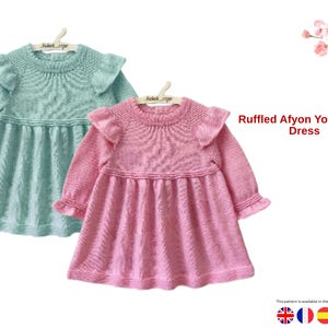 May include: Two knitted baby dresses, one in light teal and the other in pink, are displayed. Each dress features long sleeves, a ruffled yoke, and a flared skirt. The text "Ruffled Afyon Yoke Baby Dress" is visible.