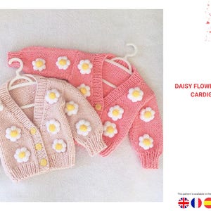Op de afbeelding: Twee roze gebreide vesten versierd met madeliefjesmotieven. Eén is lichtroze, met knopen, en de andere is donkerder roze, wikkelstijl. Beide hebben witte en gele madeliefjes applicaties. De tekst luidt "DAISY FLOWER MOTIF CARDIGAN".