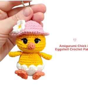 May include: A handmade yellow amigurumi chick wearing a pink hat with a white flower. The chick has black eyes, an orange beak, and orange feet. The text "Amigurumi Chick in Eggshell Crochet Pattern" is visible.