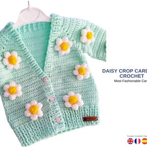 Op de afbeelding: Een lichtgroen gehaakt vest met madeliefjes. Het vest heeft een V-hals, korte mouwen en is dichtgeknoopt. De tekst op de afbeelding luidt "DAISY CROP CARDIGAN CROCHET" en "Most Fashionable Cardigan".