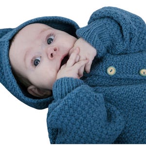 Puede incluir: Un bebé con una chaqueta con capucha de punto azul verdoso con dos botones de madera. La chaqueta tiene una capucha puntiaguda y el bebé mira hacia arriba con los ojos muy abiertos.