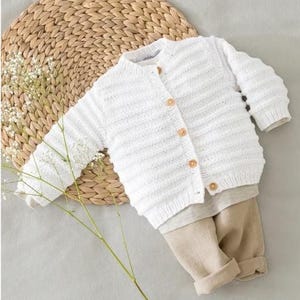 May include: White knitted baby cardigan with button closure. The cardigan is laid out on a woven straw mat with a sprig of white flowers.