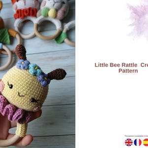 May include: A handmade crochet rattle in the shape of a bee, held in a hand. The bee is yellow with brown antennae and blue and purple flower accents. The rattle has a wooden ring base. Other crochet animal rattles are visible.