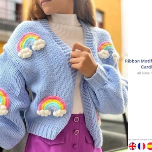 Puede incluir: Un cárdigan azul claro con frente abierto adornado con motivos de arcoíris y nubes. El suéter corto tiene cuello en V y mangas largas. El texto de la imagen dice "Ribbon Motif Adult Crop Cardigan" y enumera las tallas S-M-L-XL.