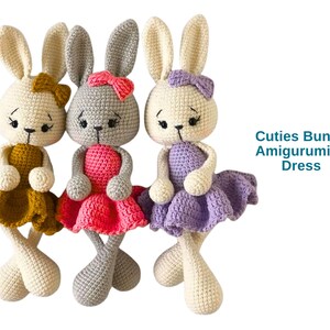 May include: Three crocheted bunny amigurumi dolls in a row, each with a different colored dress and bow. The bunnies have long ears and are in cream, gray, and cream colors. The dresses are mustard, pink, and lavender. Text reads "Cuties Bunny Amigurumi in Dress."