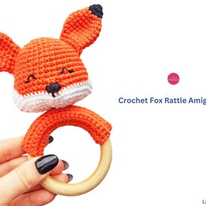 May include: Crocheted fox rattle amigurumi in orange, white, and black. The fox has large ears and a smiling face. The rattle has a wooden ring and is held in a hand. The text "Crochet Fox Rattle Amigurumi" is on the right.