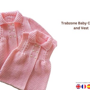 May include: A light pink knitted baby cardigan and vest set. The cardigan features a collared neckline, pearl buttons, and cable knit detailing. The vest also has pearl buttons and cable knit accents. The text "Trabzone Baby Cardigan and Vest" is visible.
