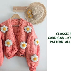 Könnte beinhalten: Ein korallenfarbener, grob gestrickter Cardigan, verziert mit großen weißen und gelben Gänseblümchen-Applikationen. Ein Strohhut mit Gänseblümchen-Akzenten hängt daneben. Der Text lautet "CLASSIC FIT CARDIGAN - KNITTING PATTERN ALL SIZES."