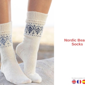 Puede incluir: Calcetines Nordic Beauty de color crema con un patrón geométrico azul en el puño. Los calcetines están tejidos y parecen estar hechos de un material suave y cálido. El texto "Nordic Beauty Socks" se muestra en la imagen.