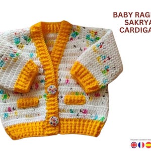 Peut inclure: Un cardigan pour bébé fait à la main, blanc avec des bordures jaunes et des motifs floraux. Le pull en crochet a un col en V, deux poches et des boutons décoratifs. Le texte "BABY RAGLAN SAKRYA CARDIGAN" est visible.