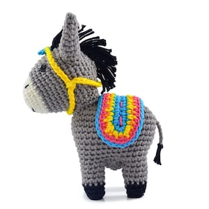 May include: A grey crocheted donkey toy with a yellow, blue, pink and green saddle. The donkey has a black mane and tail.