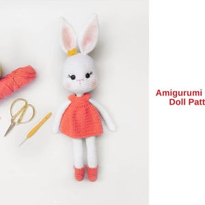 Könnte beinhalten: Eine handgefertigte weiße Amigurumi-Hasenpuppe mit einem korallenfarbenen Kleid, weißen Armen und Beinen sowie rosa gefütterten Ohren. Die Puppe befindet sich neben Garn, einer Schere und einem Häkelhaken. Der Text lautet "Amigurumi Bunny Doll Pattern."