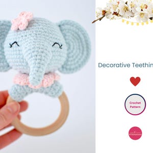 May include: A light blue crocheted elephant with pink accents attached to a wooden teething ring. The elephant has embroidered eyes and a flower on its head. The text "Decorative Teething Ring" is visible in the image.