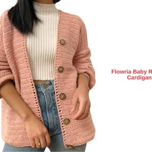 May include: A pale pink, open-front cardigan with large wooden buttons. The crochet sweater has long sleeves and is worn over a white ribbed top and blue jeans. The text "Flowria Baby Raglan Cardigan" is visible on the right side of the image.