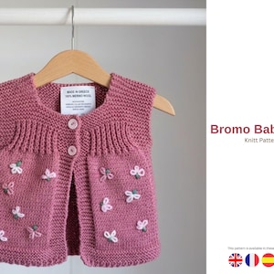 May include: A dusty rose baby vest with a floral design, hanging on a wooden hanger. The knitted vest features a round neckline, three buttons, and embroidered flowers. The text "Bromo Baby Vest" is visible.