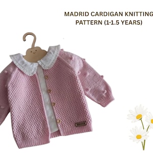 May include: A light pink knitted cardigan with a white collar and button closure, displayed on a wooden hanger. The cardigan has a textured pattern and small pom-poms on the sleeves. Text at the top reads "MADRID CARDIGAN KNITTING PATTERN (1-1.5 YEARS)".