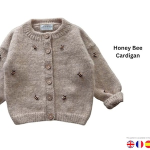 Puede incluir: Un cárdigan Honey Bee marrón claro con bordado de abejas. El suéter con botones tiene cuello redondo y mangas largas. El texto "Honey Bee Cardigan" es visible en el lado derecho de la imagen.