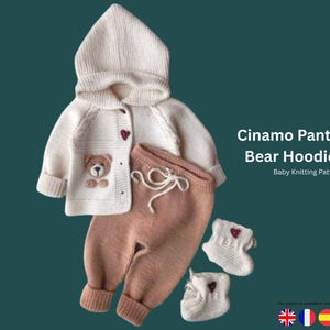 May include: A cream-coloured knitted baby set with a hooded jacket, matching trousers, and booties. The jacket has a bear applique and heart-shaped buttons. The set is in cream and brown tones. Text reads "Cinamo Pants And Bear Hoodie Set".