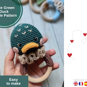 Könnte beinhalten: Gehäkelter Entenrassel mit grünem Kopf, gelbem Schnabel und beigem Kragen. Die Rassel hat einen Holzring und trägt die Aufschrift "Little Green Duck Rattle Pattern" und "Easy to make".