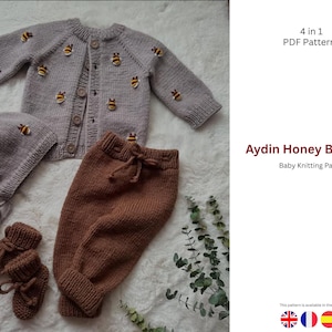 以下が含まれることがあります： グレーとブラウンのニットベビーセット。カーディガン、帽子、パンツ、ブーティーが含まれています。グレーのカーディガンと帽子には、刺繍された蜂のディテールが施されています。「Aydin Honey Baby Set」と「Baby Knitting Patterns」のテキストが含まれています。