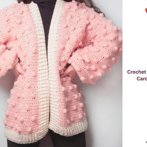 Puede incluir: Cárdigan de ganchillo rosa claro con diseño de punto de palomitas de maíz. El cárdigan abierto tiene un borde y un ribete de color crema. Las mangas son largas y la prenda está hecha a mano. El texto "Crochet Popcorn Cardigan" es visible.