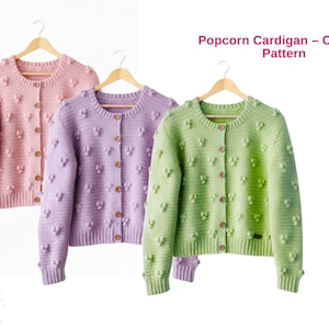 Peut inclure: Trois cardigans en crochet popcorn aux couleurs pastel : rose, lavande et vert. Chaque cardigan est boutonné et suspendu à un cintre en bois. Le texte "Popcorn Cardigan - Crochet Pattern" est affiché au-dessus des cardigans.