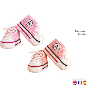 May include: Hand-crocheted baby booties in a Converse style. The pink and white shoes feature white laces, a black stripe, and a red accent. Each shoe has a white patch with a black "A" in the center. The text "Converse Style Baby Booties Crown" is visible.